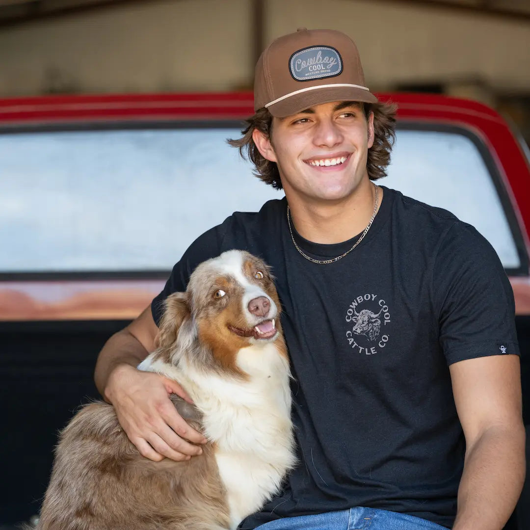 Man wearing a Cowboy Cool Lariat hat and western t-shirt, sitting on a tailgate with a dog.
