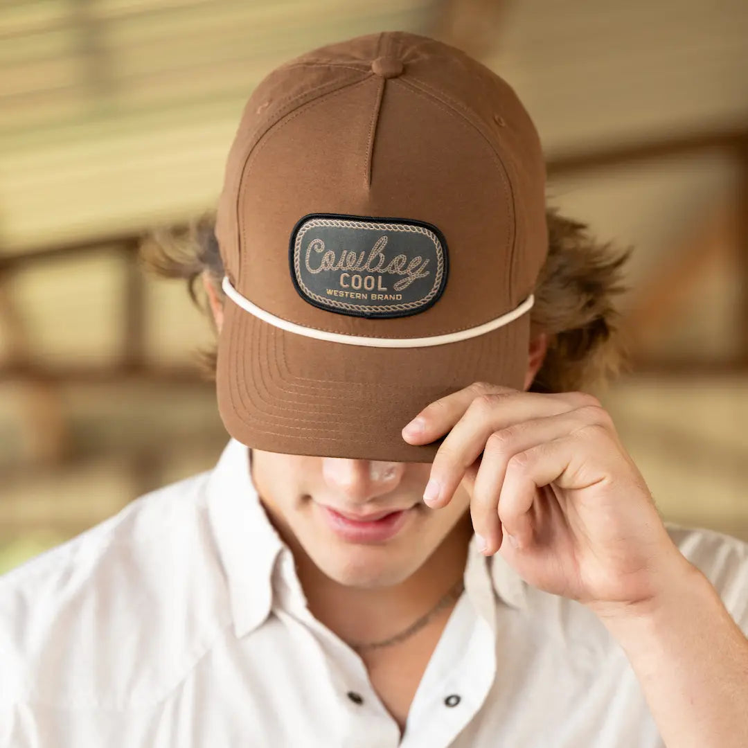 Person wearing a brown western hat with a 'Cowboy Cool' patch, holding it up to their face.