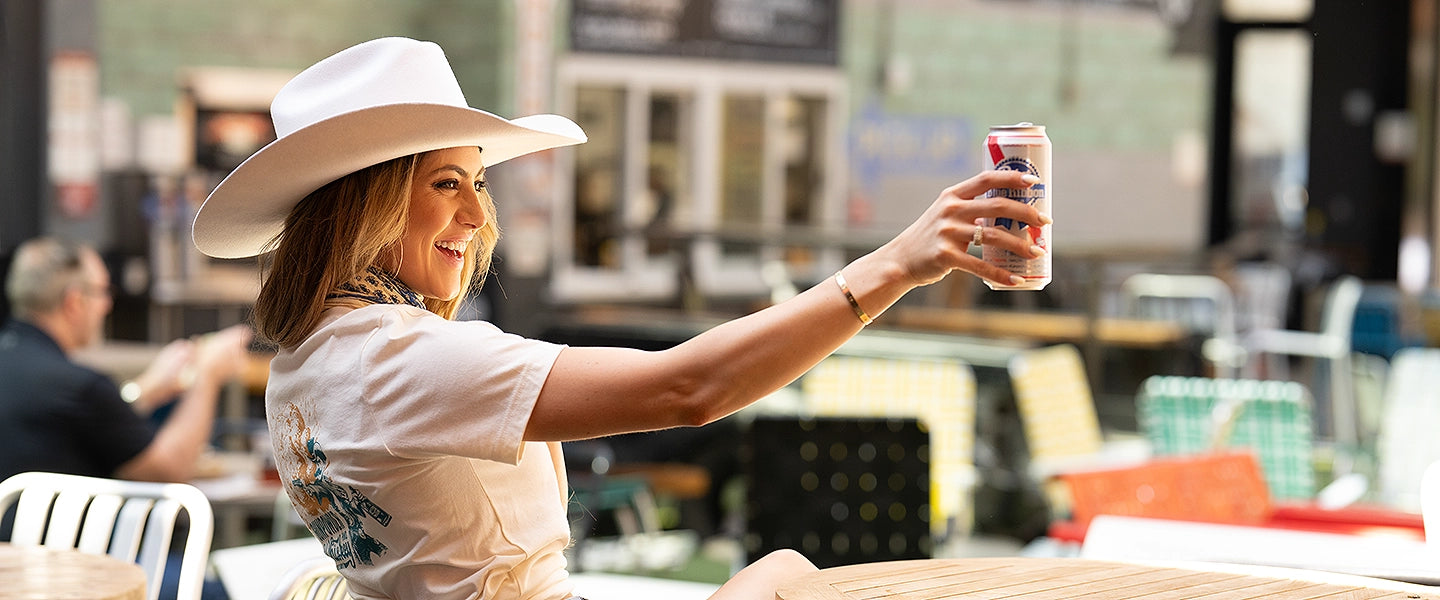 Smiling woman wearing a white cowboy hat, graphic t-shirt, denim shorts, and brown cowboy boots, raising a can of Pabst Blue Ribbon while seated outdoors. Photo for Cowboy Cool’s new arrivals collection.