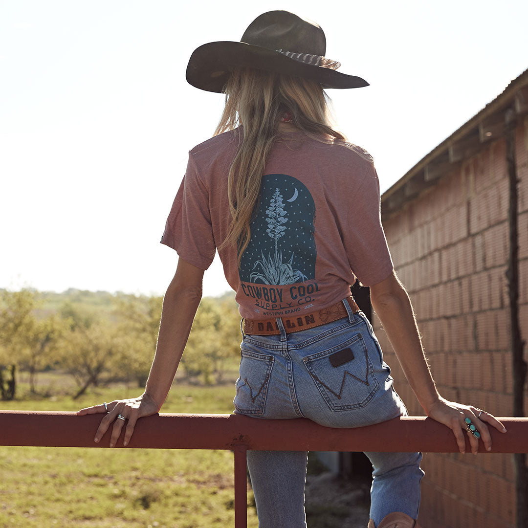 Back view of woman wearing Cowboy Cool's Century Agave T-shirt in mauve, featuring a nighttime desert scene with agave plant and crescent moon graphic. Model sits on ranch fence wearing Wrangler jeans, dark cowboy hat, and turquoise rings, with sunset-lit pasture in background. Western lifestyle photography showcasing shirt's artistic back design.