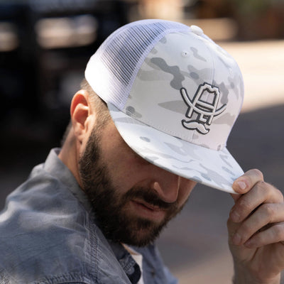 Man wearing white camo trucker hat with cowboy hat and mustache logo, adjusting brim outdoors