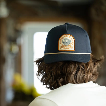 Man wearing a Cowboy Cool Dillo After Dark Hat in navy with tan rope trim and vintage style patch featuring a West Texas armadillo drinking a beer, 6 panel snapback design, facing away from the camera.