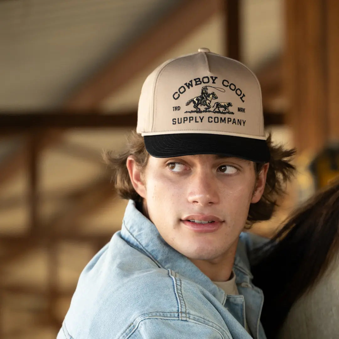 A team roper in a Cowboy Cool Roper Hat with 'Cowboy Cool Supply Company' embroidered above a calf roping design, background blurred