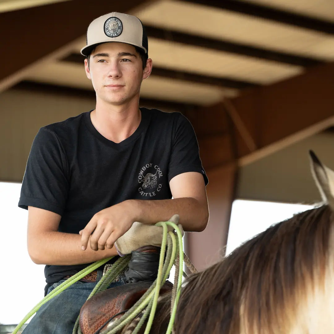Man sitting on a horse holding lariat rope, wearing a black t-shirt and  a Cowboy Cool Cattle Company hat.