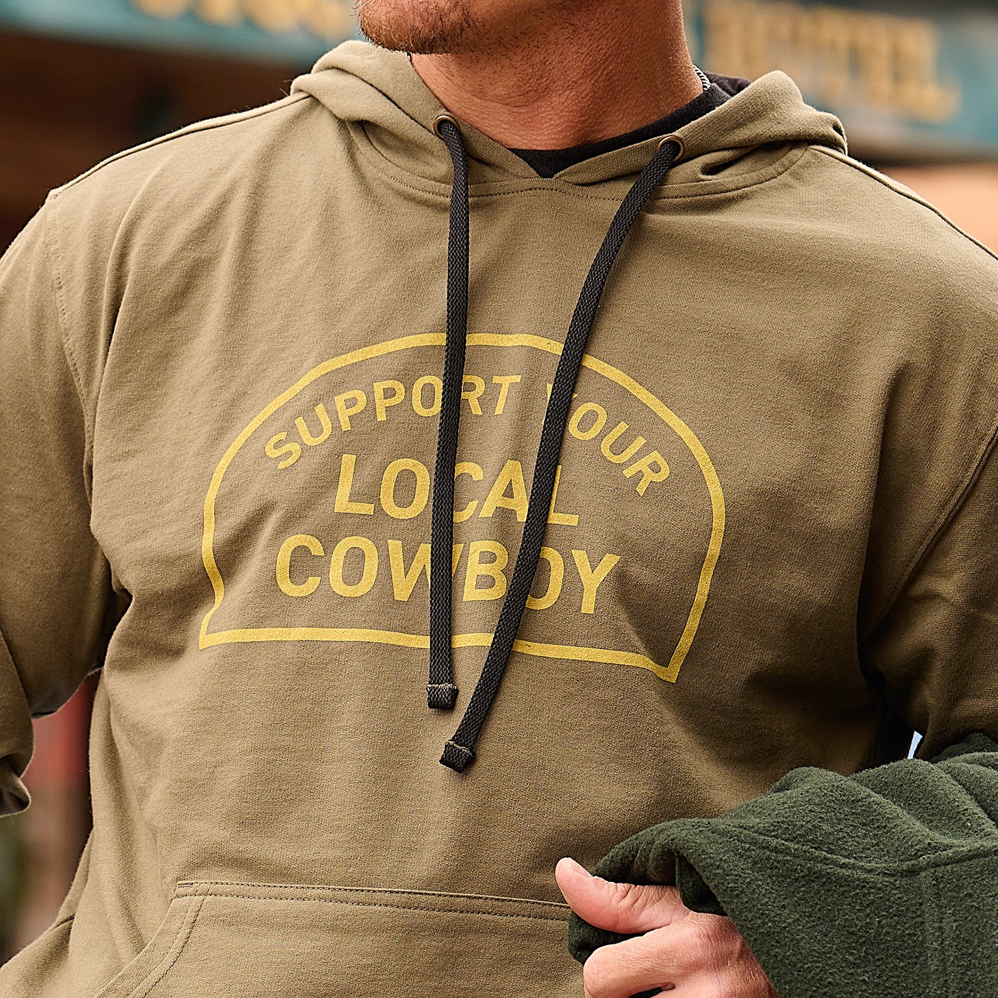 Man wearing an olive green hoodie with the text ‘Support Your Local Cowboy’ printed on the chest.