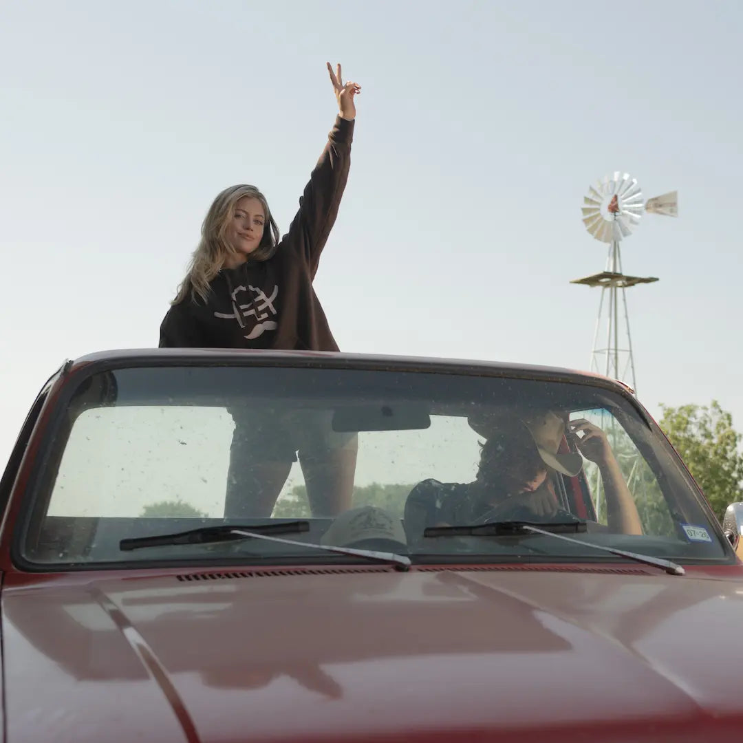 Woman standing in the back of a  truck bed wearing Cowboy Cool Hank Hoodie in brown with a windmill in the background
