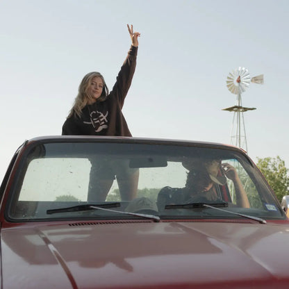 Woman standing in the back of a  truck bed wearing Cowboy Cool Hank Hoodie in brown with a windmill in the background
