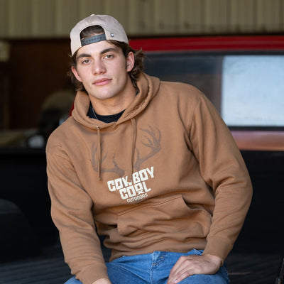 Man wearing a Brown western hoodie with 'Cowboy Cool Outdoors' graphic with a deer skull on a light beige background