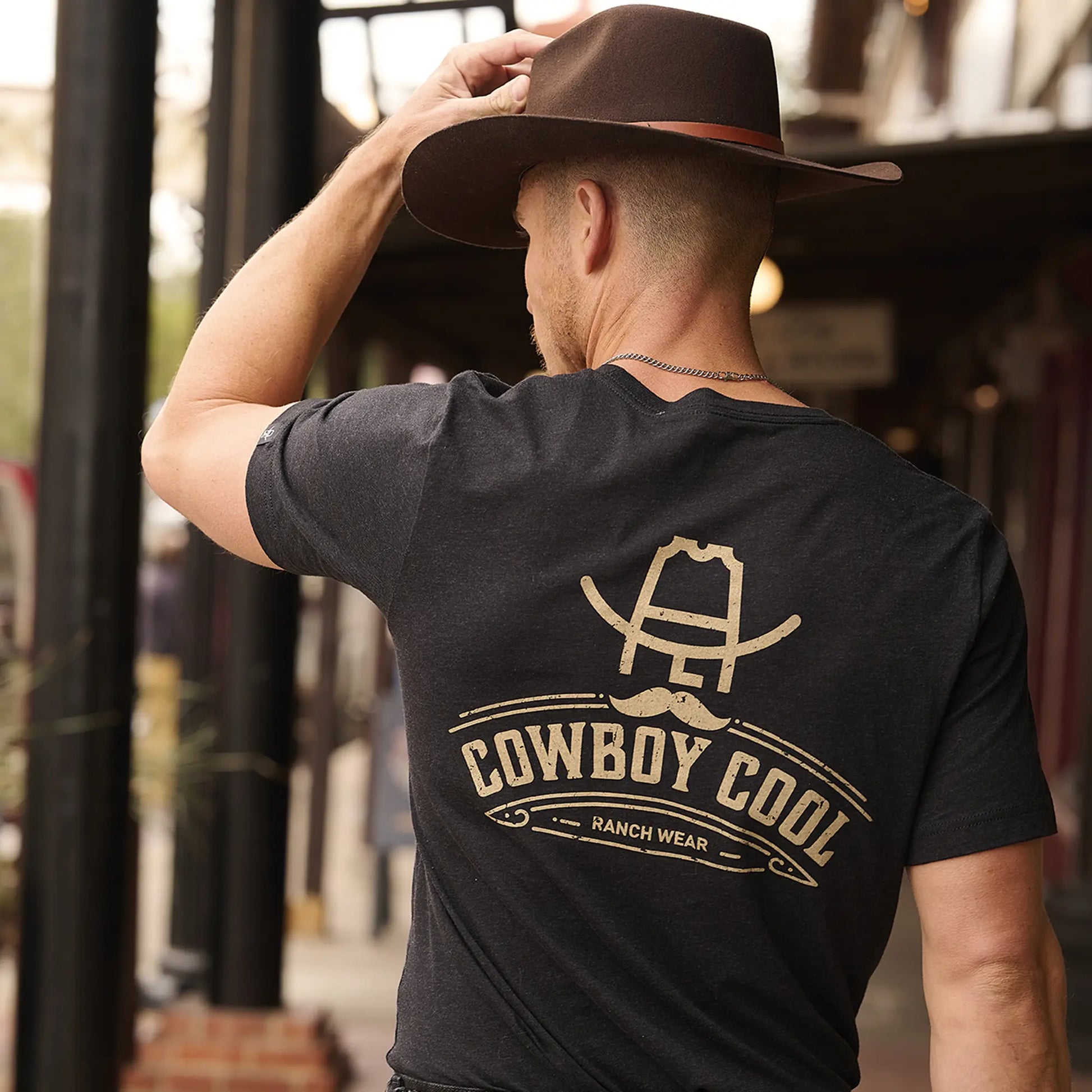 Cowboy Cool Hank Ranchwear T-Shirt shown in lifestyle setting at western bar. Classic black t-shirt features tan Cowboy Cool Ranch Wear logo with cowboy hat and western design. Model wearing shirt with western belt, jeans, and traditional cowboy hat, demonstrating versatile casual western styling. 