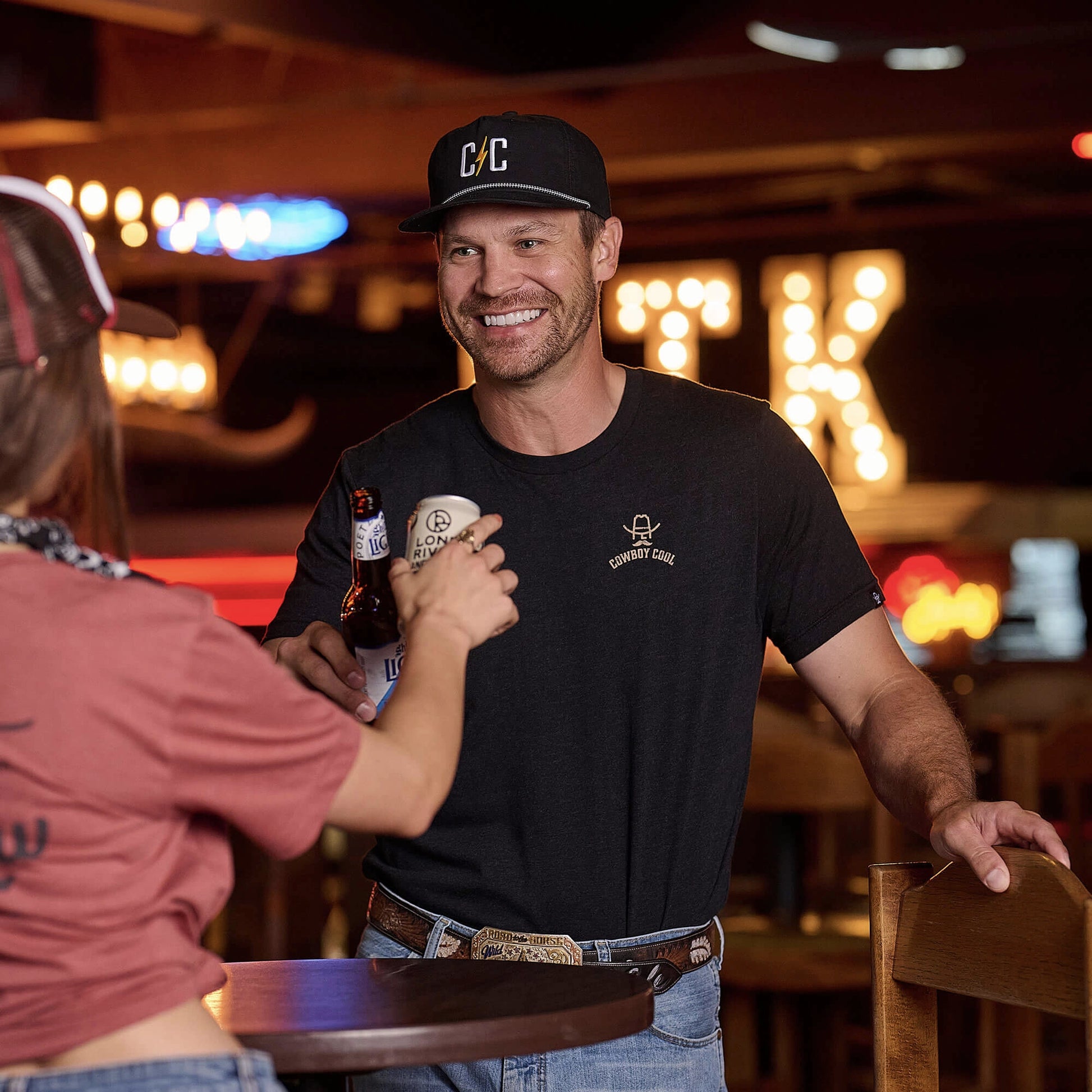 Cowboy Cool Hank Ranchwear T-Shirt shown in lifestyle setting at western bar. Classic black t-shirt features tan Cowboy Cool Ranch Wear logo with cowboy hat and western design. Model wearing shirt with western belt, jeans, and baseball cap, demonstrating versatile casual western styling. 