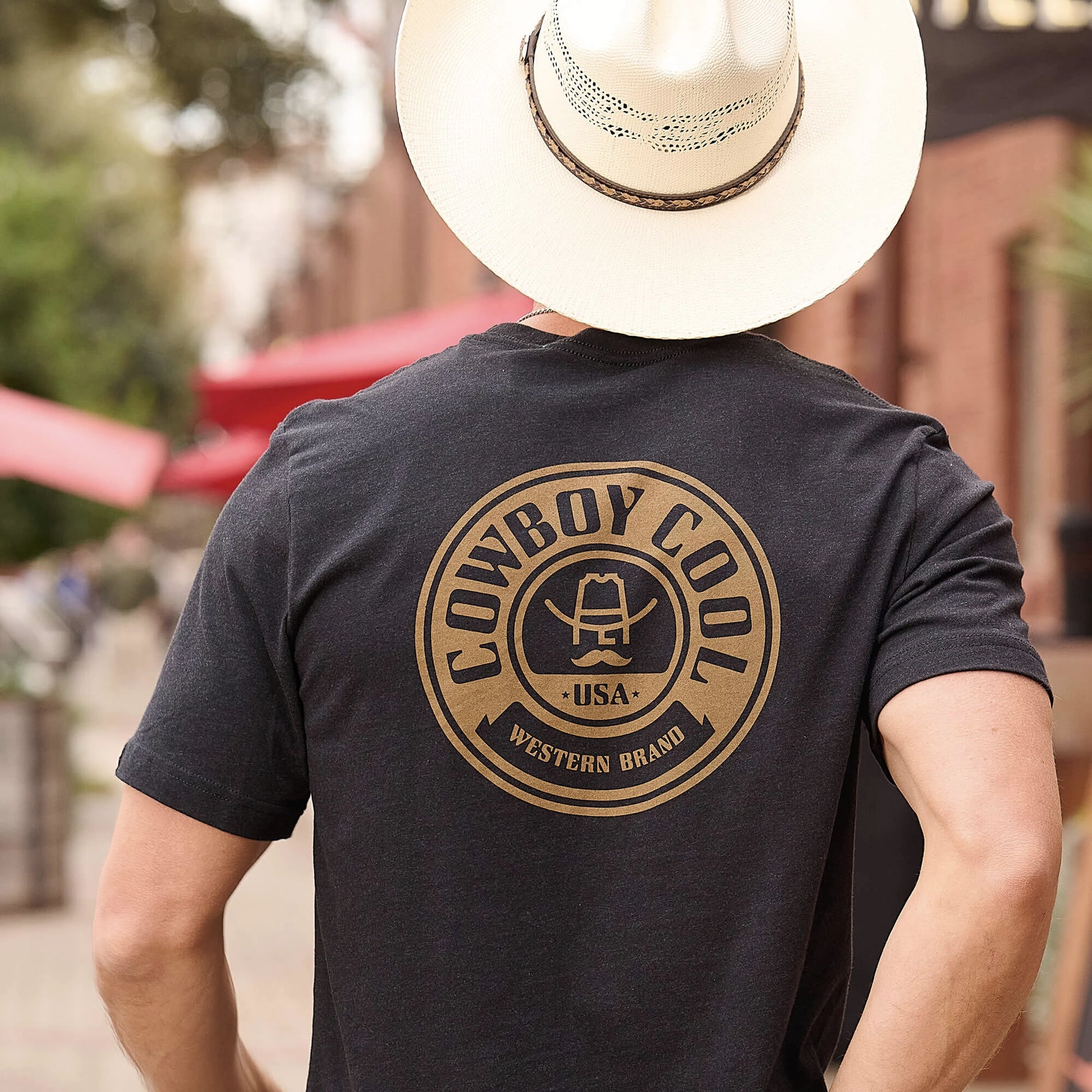 A man wearing a black Maverick T-shirt with an emblem on the back, paired with a tan cowboy hat. 