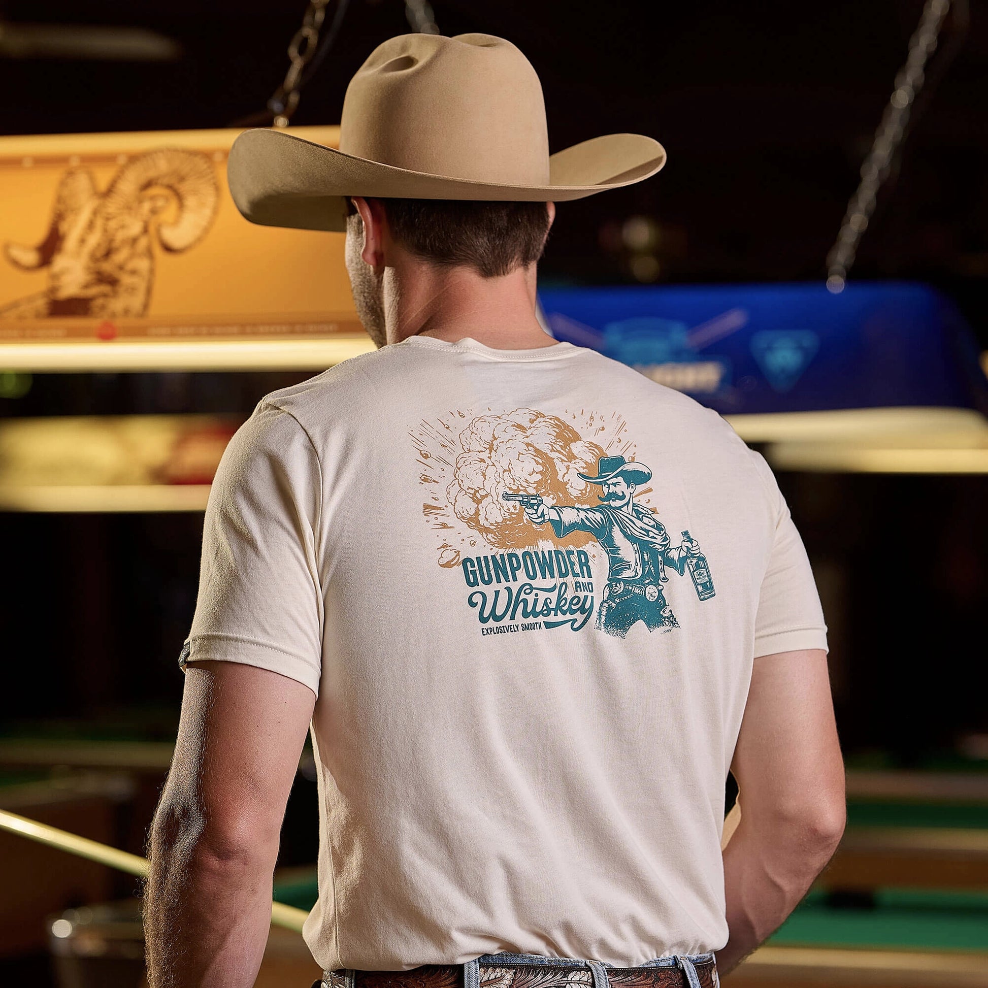 Back view of Gunpowder & Whiskey t-shirt in cream, featuring large vintage-style graphic of cowboy with pistol and whiskey bottle in teal and bronze tones. Model wearing tan cowboy hat and leather belt, photographed in bar setting with pool table visible in background.