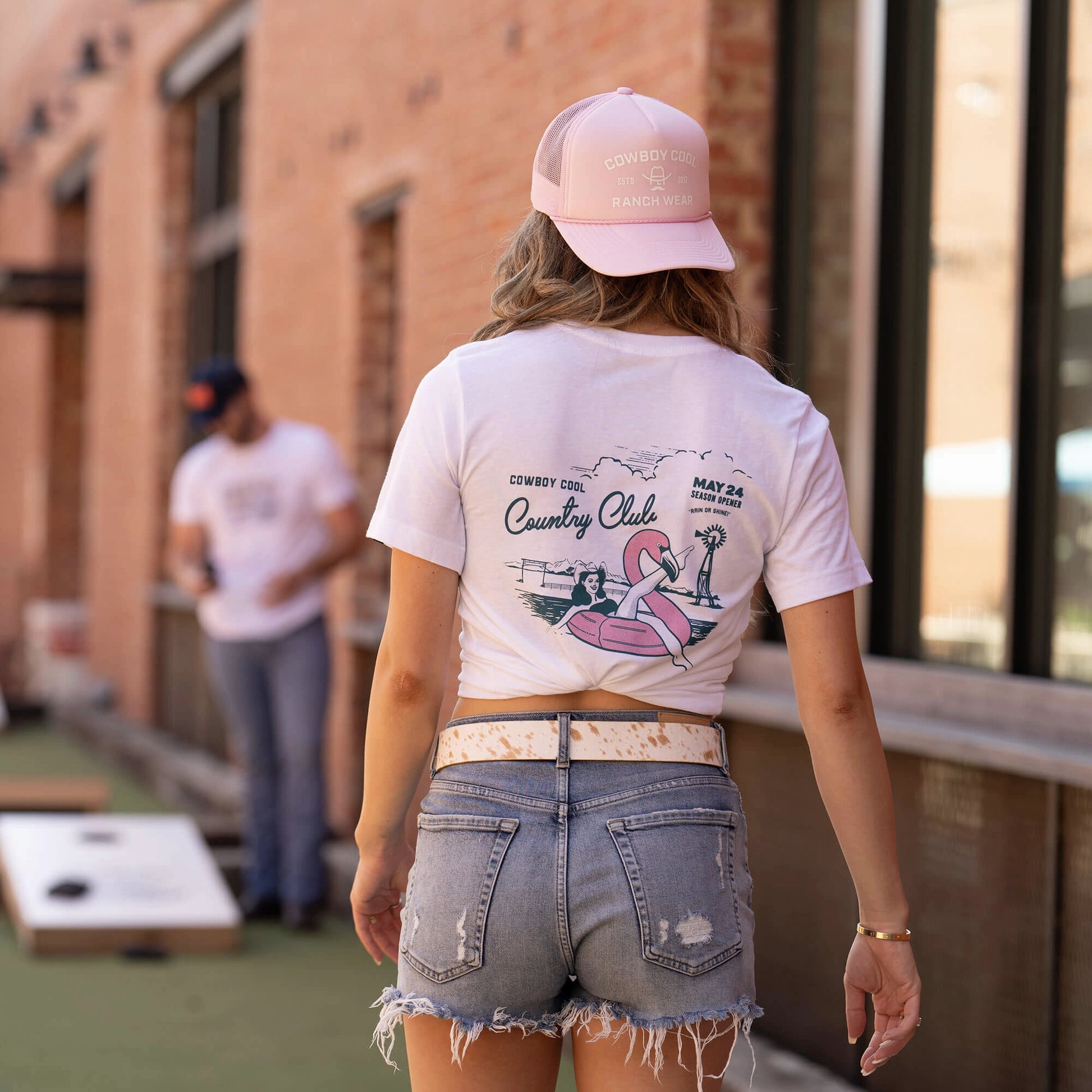 Back view of woman wearing Cowboy Cool's Country Club T-shirt in white, featuring a playful western-meets-pool-party graphic design with pink flamingo float and 'May 24 Season Opening' text. Shirt is styled casually knotted, paired with distressed denim shorts and cowhide print belt. Model also wears matching pink Cowboy Cool Ranch Wear trucker hat. Casual outdoor setting with cornhole game visible in background. Western lifestyle street wear photography showcasing shirt's summer-themed back design.